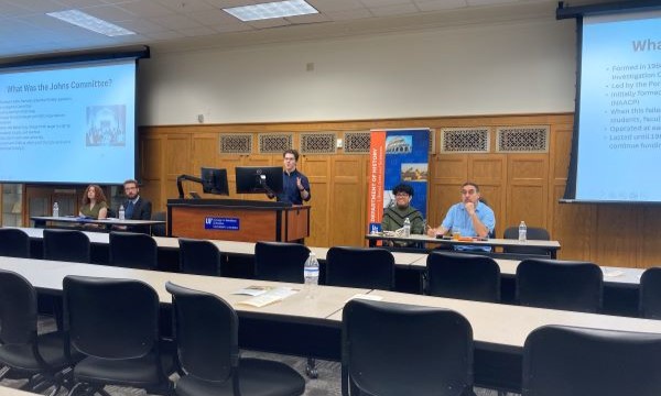 a group of people in a meeting in a conference hall. They are seated at a table with chairs, and there is a presentation being displayed on a screen.