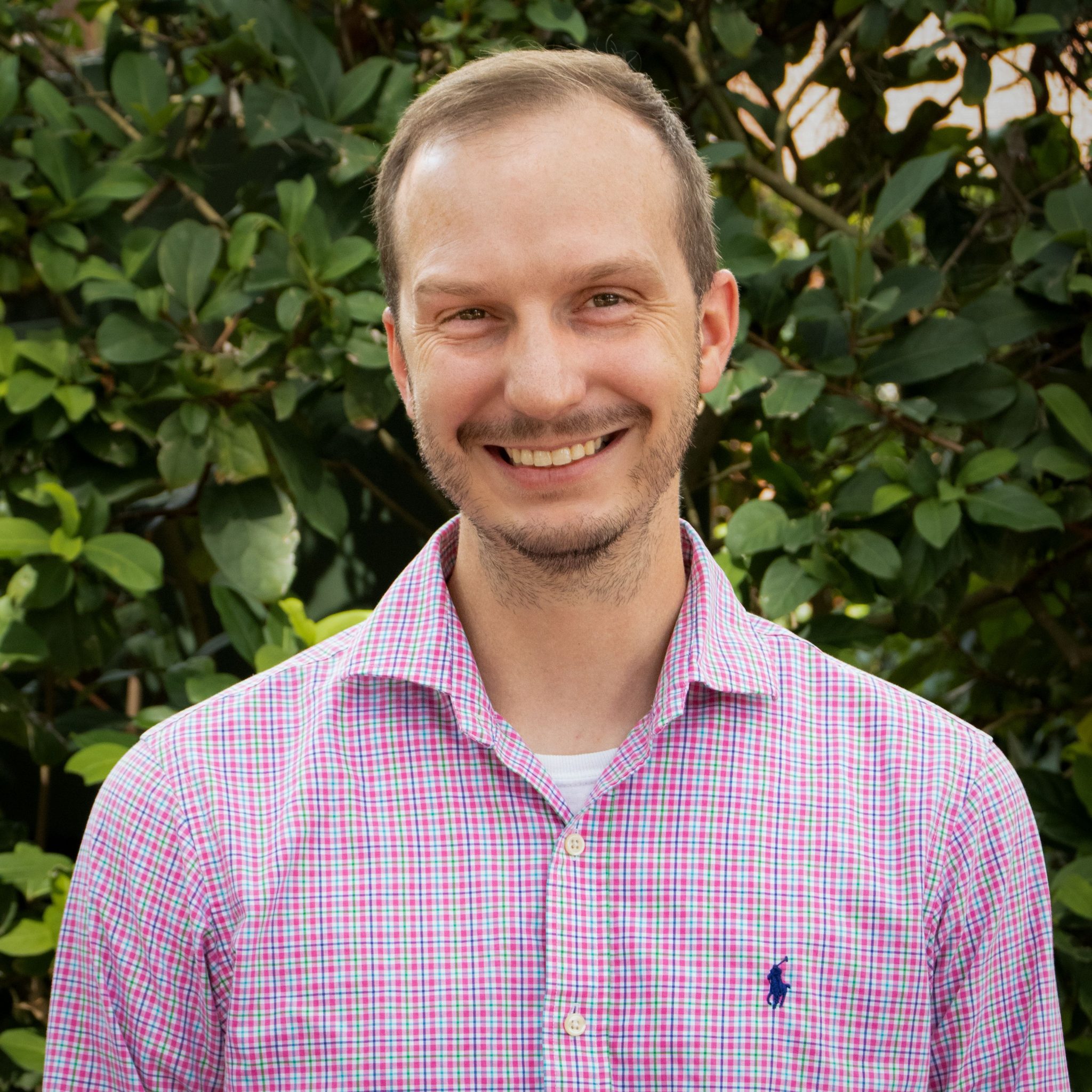 Smiling man with a short beard wearing a pink, green, and white plaid shirt, standing in front of green foliage.